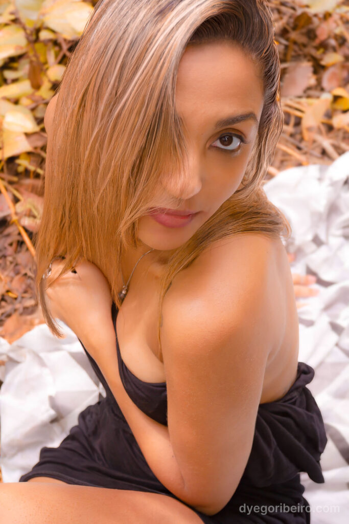 Modelo em ensaio nu artístico na natureza em Niterói, posando de forma sensual com blusa preta caída no ombro e folhas secas ao fundo.