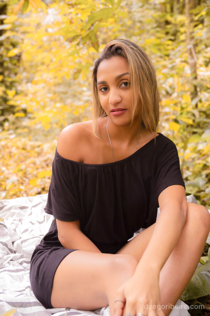 Início de ensaio nu artístico em Niterói com mulher sentada na natureza usando blusa preta caída nos ombros.