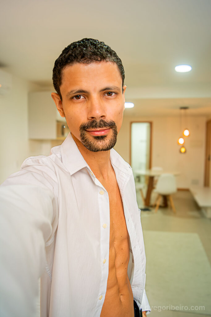 Retrato autoral do fotógrafo Dyego Ribeiro com camisa branca aberta, em ambiente interno com iluminação quente.
