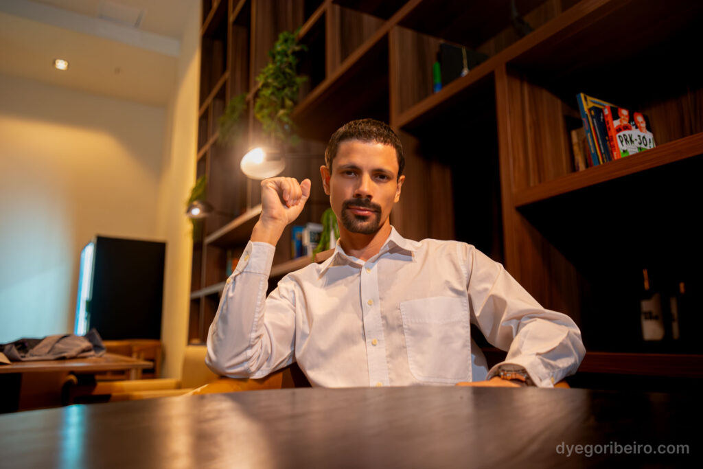 Dyego Ribeiro sentado à mesa em seu autorretrato profissional, evidenciando o uso de iluminação natural para ensaios femininos em Niterói.