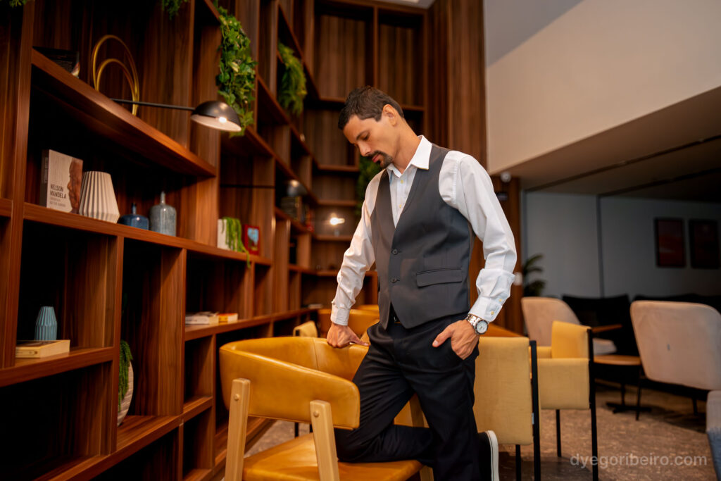 Retrato corporativo de Dyego Ribeiro olhando para baixo enquanto ajusta o colete em ambiente decorado com estantes de madeira.