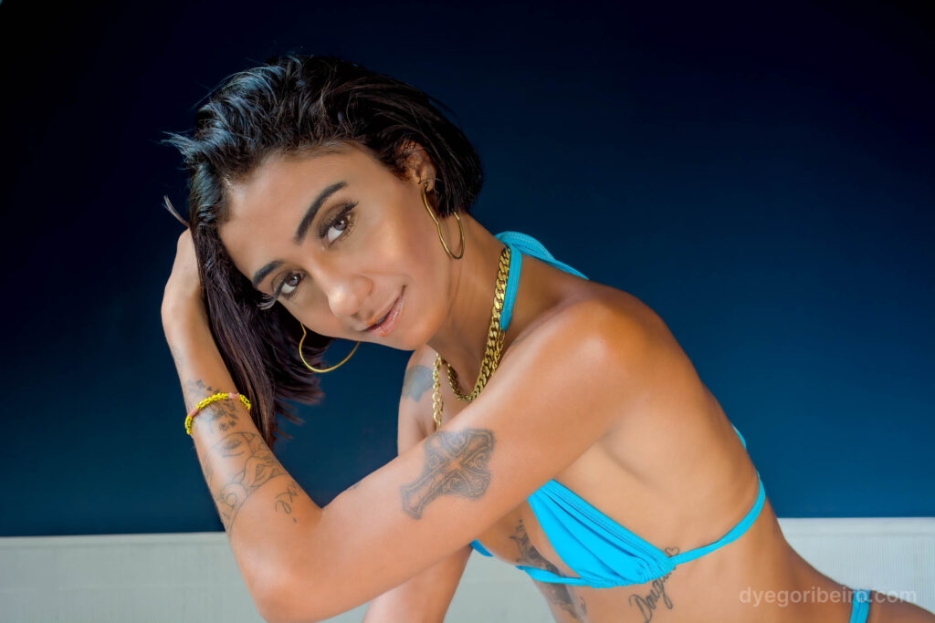 Criadora de conteúdo olhando para a câmera, ajoelhada na cama com calcinha e biquini azul, posando para um ensaio sensual Niterói para o CloseFans.