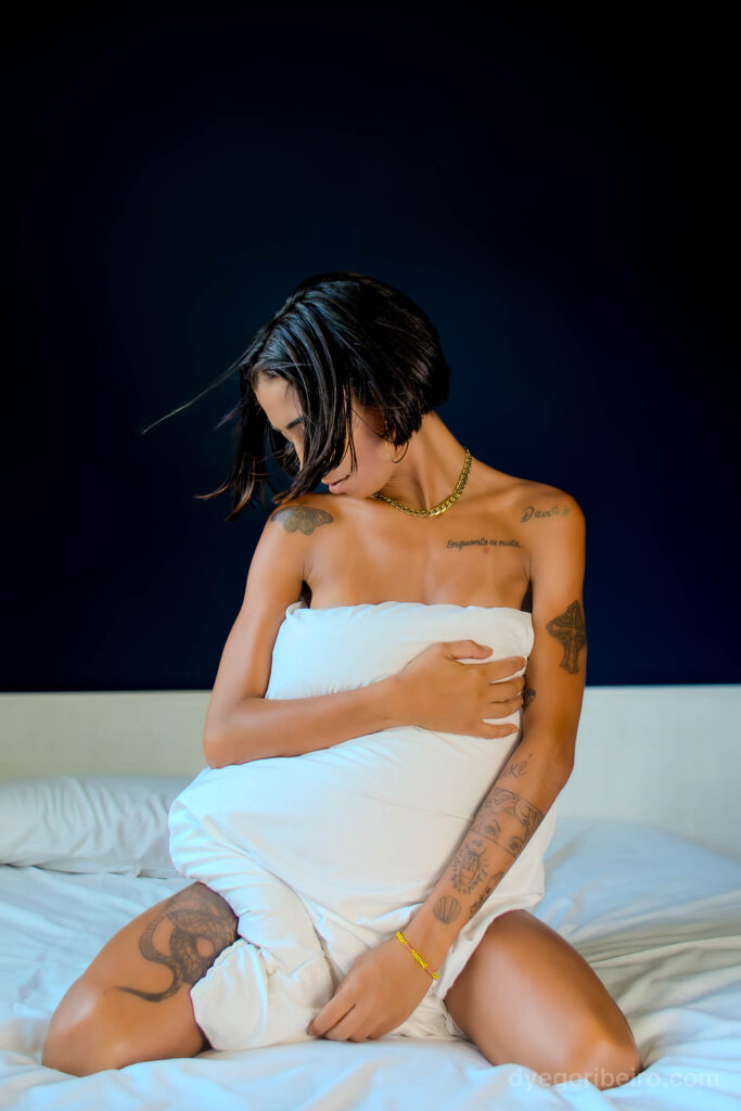 Mulher sentada na cama se cobrindo levemente com lençóis brancos e um travesseiro, em uma vibe de ensaio sensual Niterói intimista.